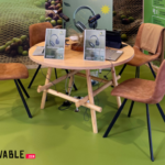 una mesa con cuatro sillas alrededor con dos tablets y dos microfonos sobre la mesa en un stand de feria ENOMAQ 2025